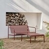 HAY Palissade Gartenbank aus Metall in Rot auf gepflastertem Weg vor weißer Wand und Holzstapel - Palissade Gartenbeistelltisch